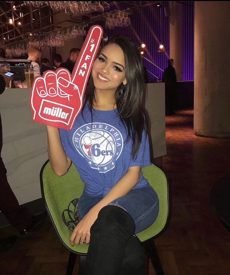 Lisa Ramos smiling while sitting in a chair, wearing a blue Philadelphia 76ers t-shirt and holding a red foam finger that says '#1 FAN'.
