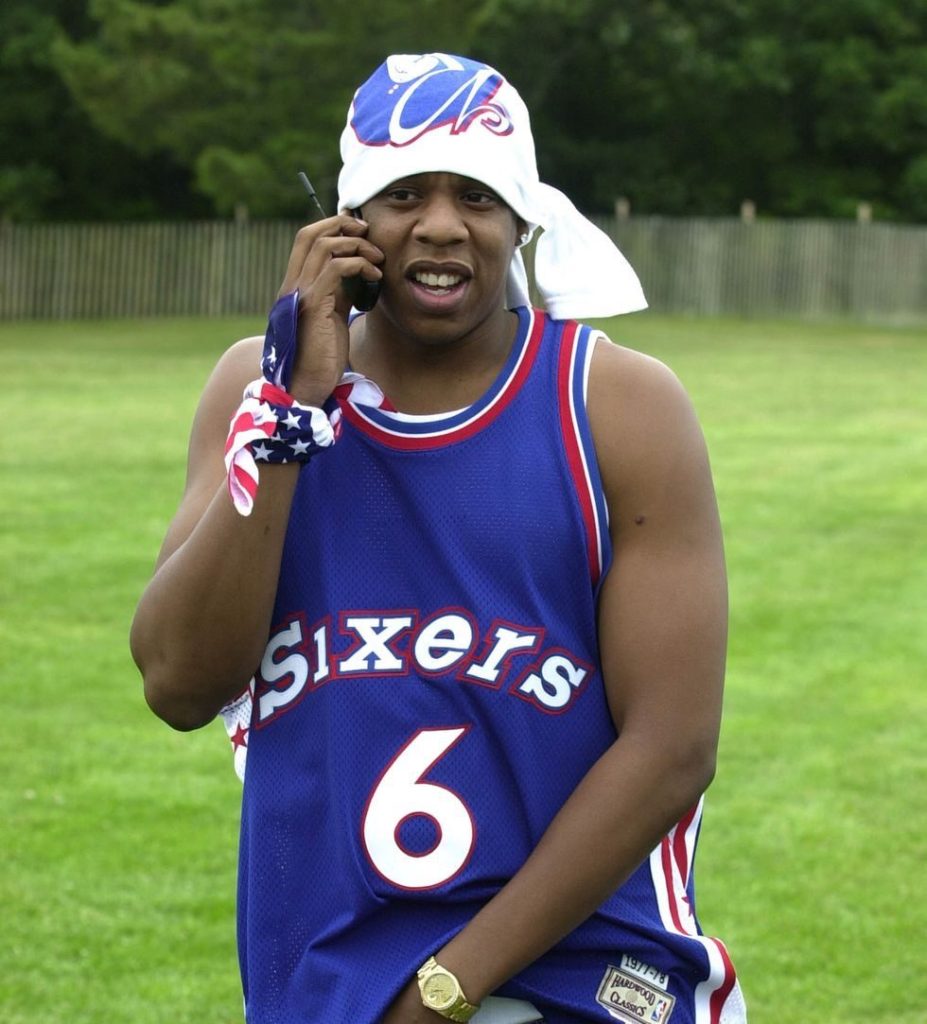Jay-Z talking on a cellphone while wearing a blue Philadelphia Sixers jersey with the number 6, standing in a grassy area.