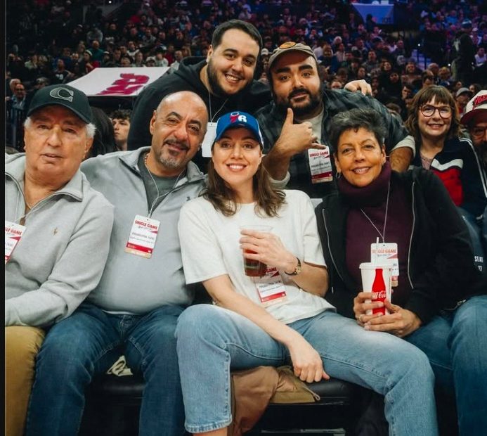 Aubrey Plaza and her family sitting together at a basketball game, smiling and enjoying their time. The woman in the front is wearing a blue cap and holding a drink, while the others are casually dressed and seated behind her.