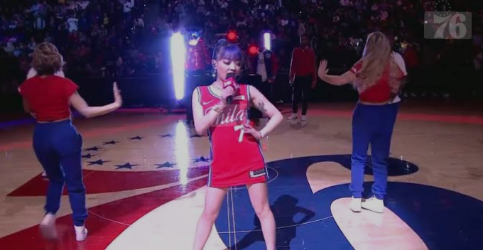 Chinese rapper VaVa performing at a Philadelphia 76ers basketball game, wearing a red Sixers jersey with the number 7. She is holding a microphone and surrounded by dancers in the background.