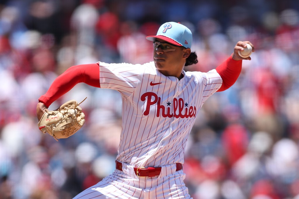 A baseball player in a red and white striped uniform with 'Phillies' on the chest is seen pitching on a sunny day. The player wears a light blue cap and is in the middle of a throwing motion.